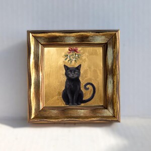 May include: A square painting in a distressed gold frame featuring a black cat sitting centered against a textured gold leaf background. Above the cat, a sprig of mistletoe with white berries and a red ribbon bow is painted in a detailed style.