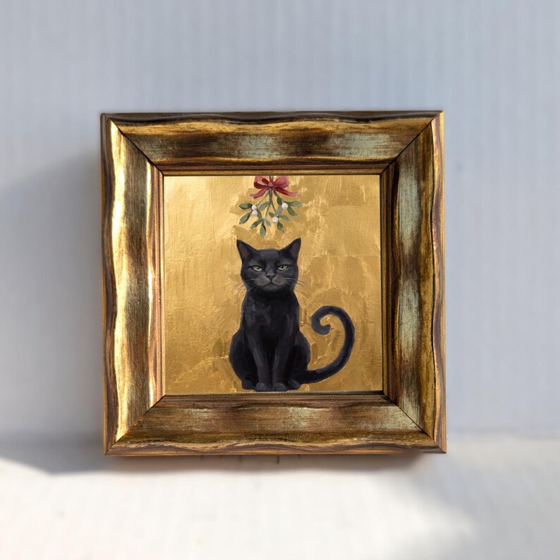 May include: A square painting in a distressed gold frame featuring a black cat sitting centered against a textured gold leaf background. Above the cat, a sprig of mistletoe with white berries and a red ribbon bow is painted in a detailed style.