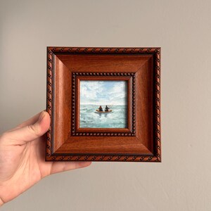 May include: A small framed painting depicting two figures in a boat on a blue ocean under a cloudy sky. The painting is in a square, dark brown wooden frame with a beaded inner border. The artwork is held by a hand.