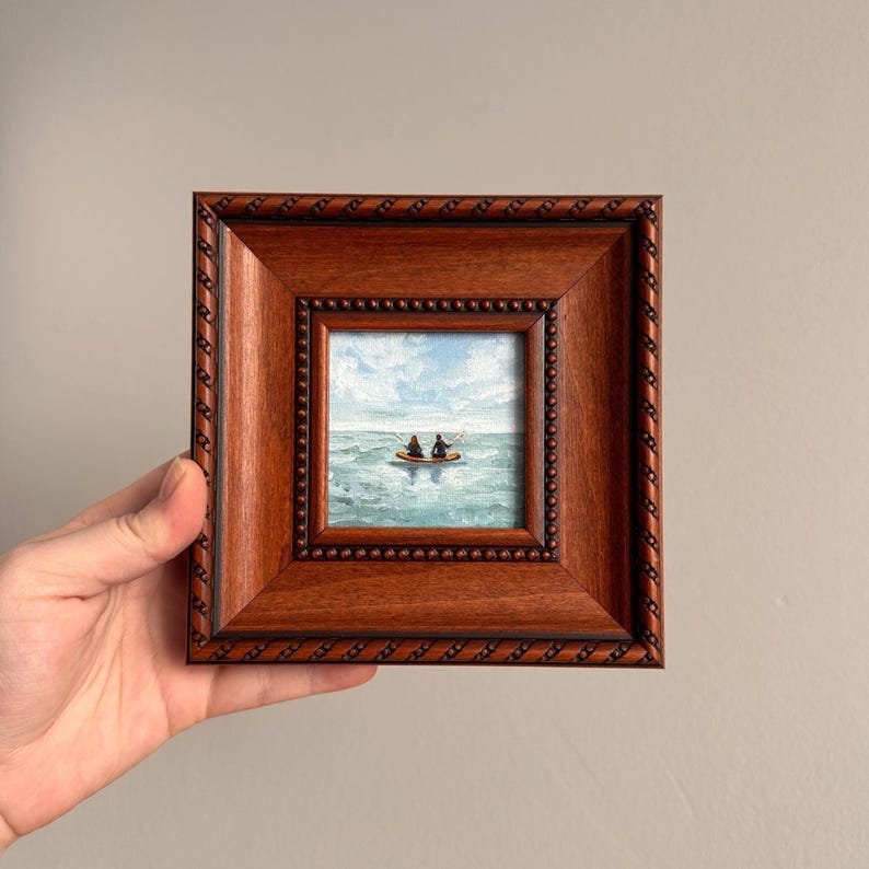 May include: A small framed painting depicting two figures in a boat on a blue ocean under a cloudy sky. The painting is in a square, dark brown wooden frame with a beaded inner border. The artwork is held by a hand.