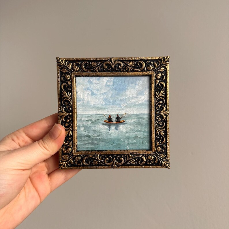 May include: A small, square painting in a decorative gold frame. The artwork depicts two figures in a boat on a blue-green sea under a cloudy sky. The frame has an ornate, vintage design, adding a touch of elegance to the seascape.