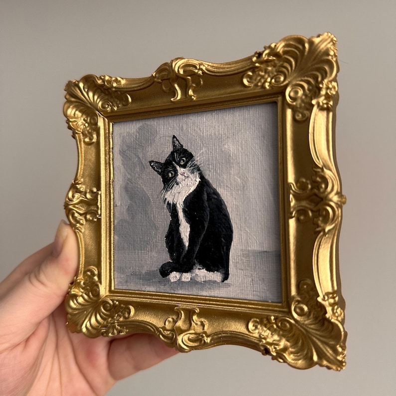 May include: A small, framed painting of a black and white cat with a curious expression. The painting is set in a gold-colored, ornate frame. The cat is depicted against a gray background.