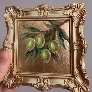 May include: A small, square painting of green olives on a branch with leaves, set in a gold ornate frame. The background is a textured gold. The painting is held by a hand.