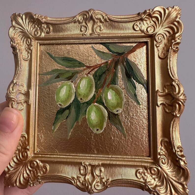 May include: A small, square painting of green olives on a branch with leaves, set in a gold ornate frame. The background is a textured gold. The painting is held by a hand.