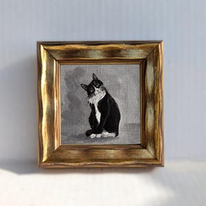 May include: A framed painting of a black and white cat with a gold-colored ornate frame. The cat is sitting and looking towards the viewer. The background is a muted gray color.