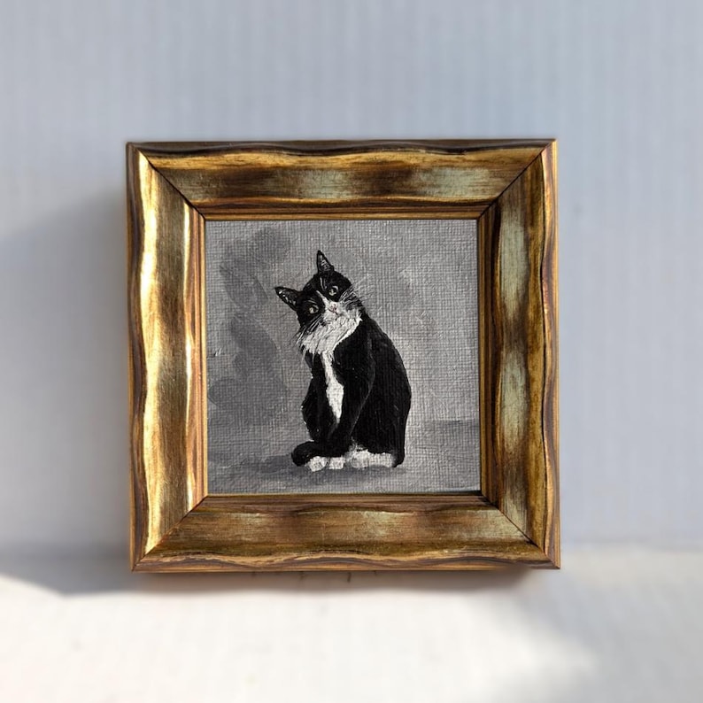 May include: A framed painting of a black and white cat with a gold-colored ornate frame. The cat is sitting and looking towards the viewer. The background is a muted gray color.