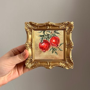 May include: A small, square, gold-framed painting featuring two vibrant red pomegranates on a branch with green leaves. The background is a textured gold surface. The ornate frame adds a touch of elegance to the artwork.