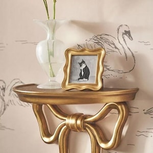 May include: A gold-colored wall shelf with a bow design, holding a small framed picture of a black and white cat. A clear vase with orange flowers sits on the shelf. The background features a swan pattern.