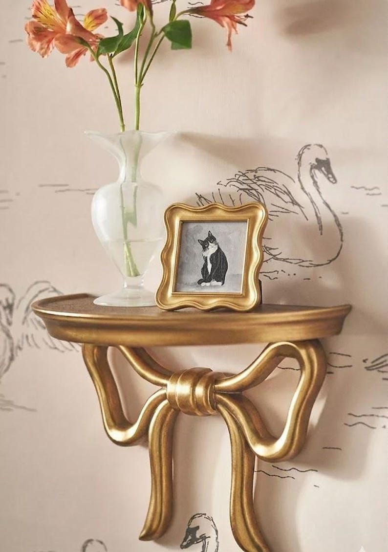 May include: A gold-colored wall shelf with a bow design, holding a small framed picture of a black and white cat. A clear vase with orange flowers sits on the shelf. The background features a swan pattern.