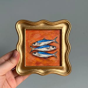 May include: A framed painting of three blue and white fish on an orange background. The artwork is in a gold-colored, ornate frame. The painting has a textured, oil-paint appearance.