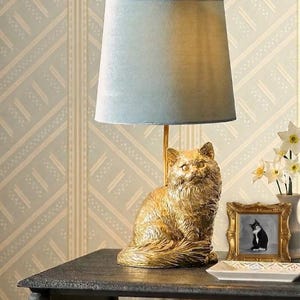 May include: A gold cat-shaped table lamp with a light blue lampshade. The lamp sits on a dark table next to a framed picture of a cat and a vase of flowers. Books are stacked below the table.