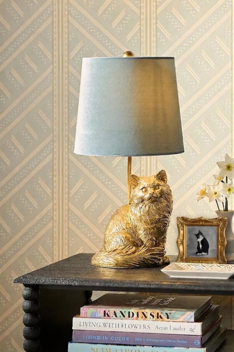 May include: A gold cat-shaped table lamp with a light blue lampshade. The lamp sits on a dark table next to a framed picture of a cat and a vase of flowers. Books are stacked below the table.