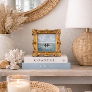 May include: A small framed painting with a gold ornate frame depicts a boat on the water. The painting rests on a stack of books with the words "Coastal" and "At Home" printed on them. A lit candle and decorative items complete the coastal theme.