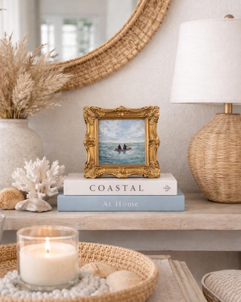 May include: A small framed painting with a gold ornate frame depicts a boat on the water. The painting rests on a stack of books with the words "Coastal" and "At Home" printed on them. A lit candle and decorative items complete the coastal theme.