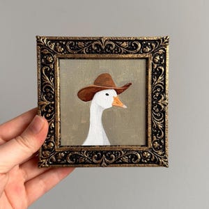 May include: A framed painting of a white goose wearing a brown cowboy hat. The artwork is set against a muted green background and framed in an ornate, gold-colored frame. The painting is held by a hand.