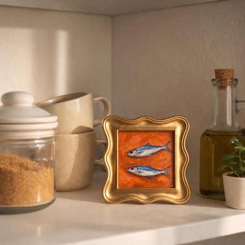May include: A small, framed painting of two fish with blue and silver details against an orange background. The painting is in a gold, ornate frame. The image is on a white shelf with other kitchen items, including a glass jar, mugs, and a bottle of olive oil.