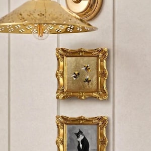 May include: A gold-toned wall sconce with a perforated shade and two small framed paintings. One painting depicts three bees on a gold background, and the other shows a black and white cat. The frames have ornate detailing.