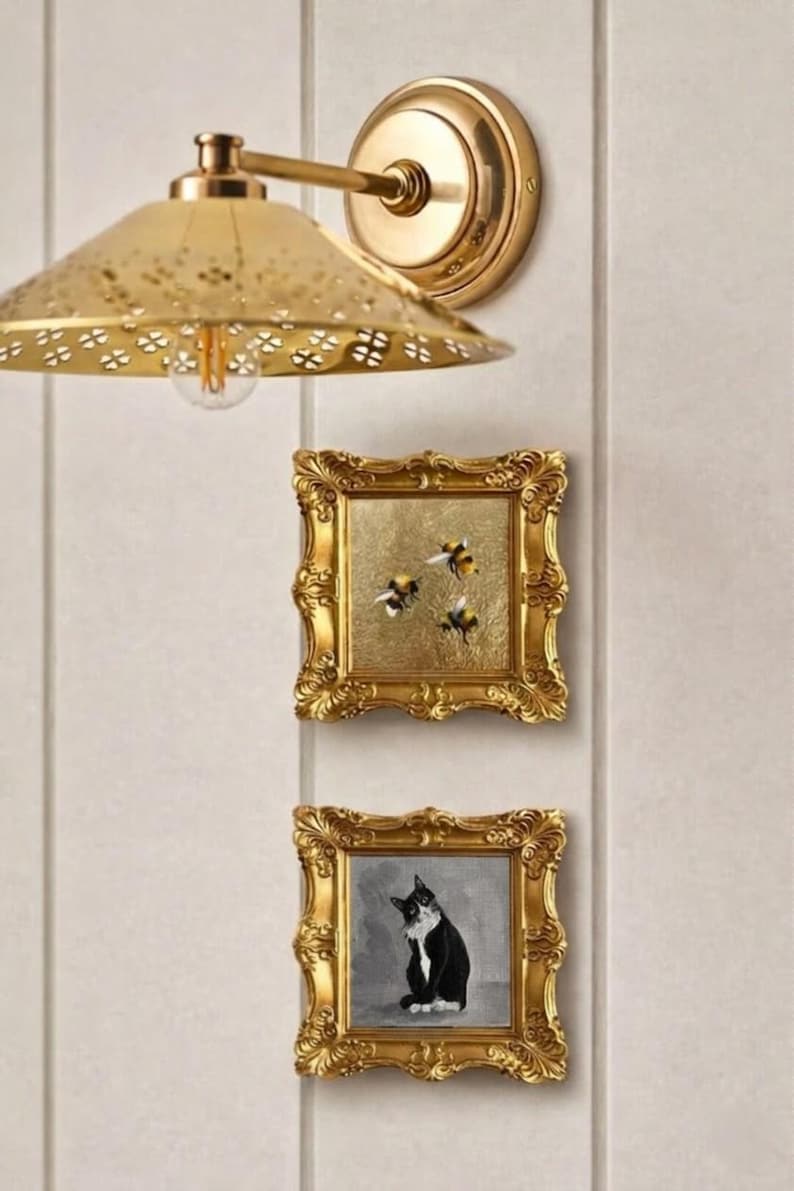May include: A gold-toned wall sconce with a perforated shade and two small framed paintings. One painting depicts three bees on a gold background, and the other shows a black and white cat. The frames have ornate detailing.