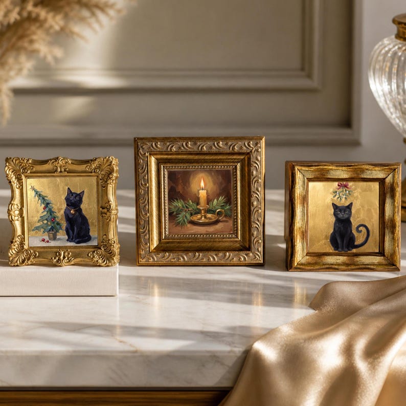 May include: Three square, ornate gold frames containing holiday-themed artwork. The left frame shows a black cat beside a small Christmas tree. The center frame depicts a lit candle in a holder with pine branches. The right frame features a black cat.