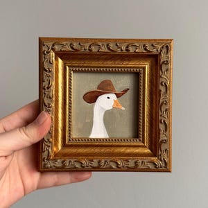 May include: A framed painting of a white goose wearing a brown cowboy hat. The artwork is set within a gold-colored, ornate frame. The goose has an orange beak and is set against a muted green background.