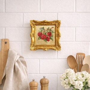 May include: A small, square framed painting with a gold ornate frame. The painting depicts red raspberries on a branch with green leaves against a gold background. The painting is hanging on a white brick wall.