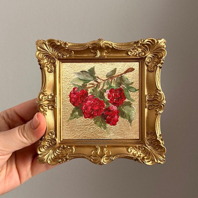 May include: A small, gold-coloured ornate frame displays a painting of red raspberries on a branch with green leaves. The painting is set against a gold textured background. The frame has a detailed, vintage design.
