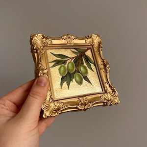 May include: A small, square painting of green olives on a branch with leaves, set in a gold ornate frame. The painting has a gold background and is held by a hand. The frame has a detailed, vintage design.