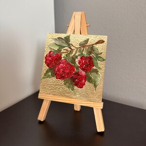 May include: Miniature painting of red raspberries on a gold background, displayed on a small wooden easel. The artwork features detailed brushstrokes and green leaves, creating a realistic depiction of the fruit. The easel is made of light-colored wood.