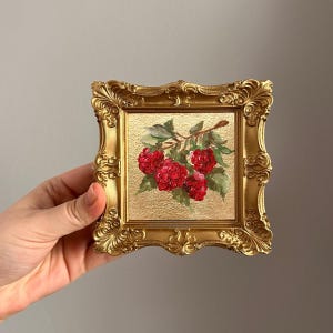 May include: A small, square, gold-framed painting featuring a cluster of vibrant red raspberries on a branch with green leaves. The painting has a textured gold background. The ornate frame adds a touch of elegance to the artwork.