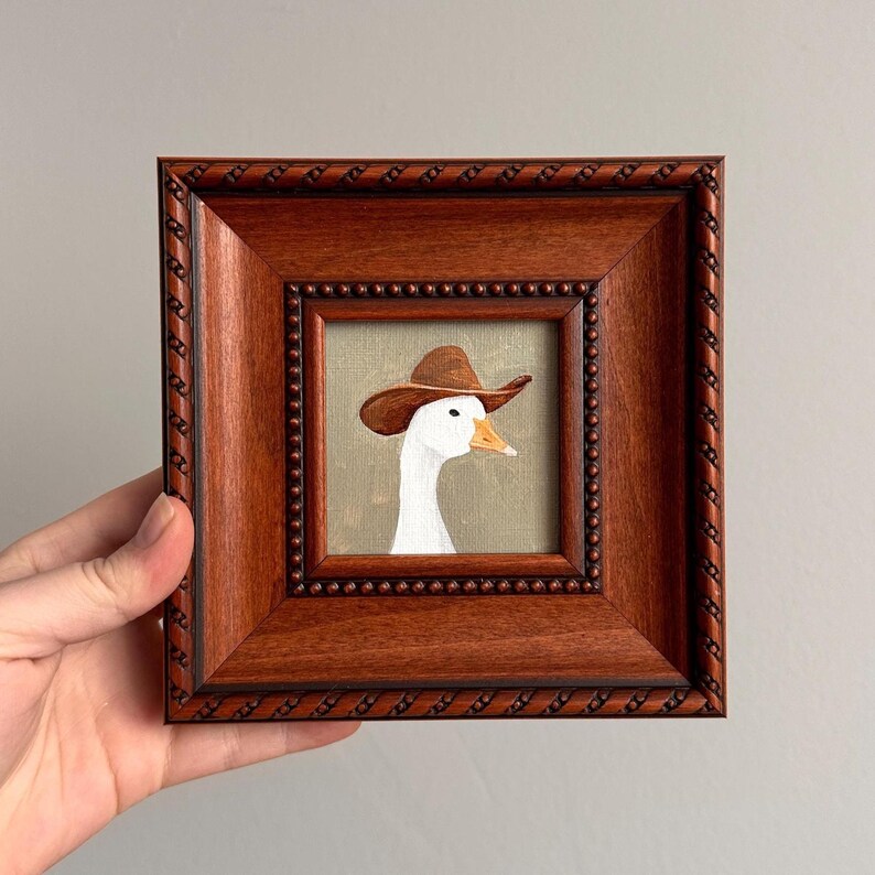 May include: A framed painting of a white goose wearing a brown cowboy hat. The artwork is set against a muted green background and encased in a dark brown, ornate wooden frame. The frame has a beaded inner border and a rope-like outer edge.
