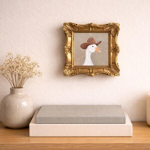 May include: A framed artwork featuring a white goose wearing a brown cowboy hat. The gold ornate frame hangs on a cream-colored wall. A vase with dried flowers and two stacked boxes sit on a wooden surface.