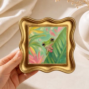 K&ouml;nnte beinhalten: Ein kleines quadratisches Gem&auml;lde eines gr&uuml;nen Frosches, der hinter einem gro&szlig;en Blatt hervorlugt, in einem dekorativen goldenen Rahmen mit gewellten Kanten. Das Kunstwerk zeigt lebhafte Pinselstriche in Gr&uuml;n, Rosa und Gelb.