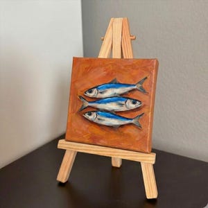 May include: A small, square painting on a wooden easel. The painting features three blue and silver fish against an orange background. The easel is made of light-colored wood.