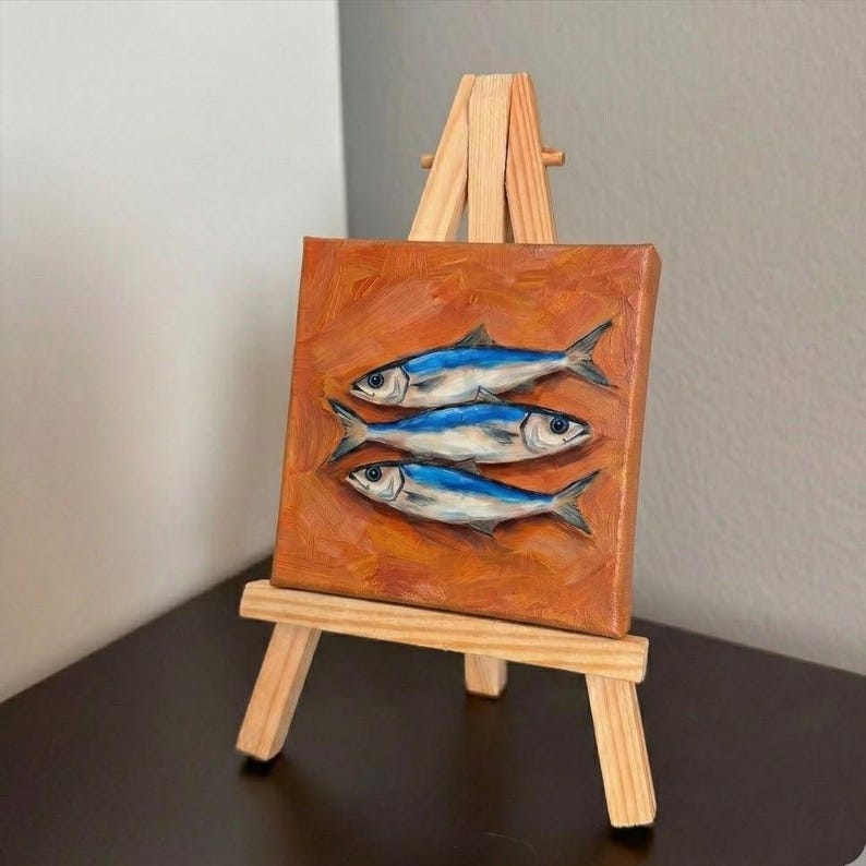 May include: A small, square painting on a wooden easel. The painting features three blue and silver fish against an orange background. The easel is made of light-colored wood.