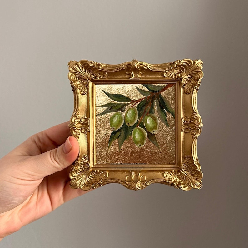May include: A small, square, gold-colored framed painting featuring a branch of green olives and leaves. The painting has a gold background and is held by a hand. The frame has ornate detailing.