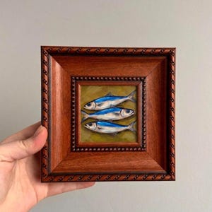 May include: A framed painting featuring three blue and silver fish on a yellow-green background. The artwork is encased in a square, dark brown wooden frame with a decorative border. The painting is held by a hand.