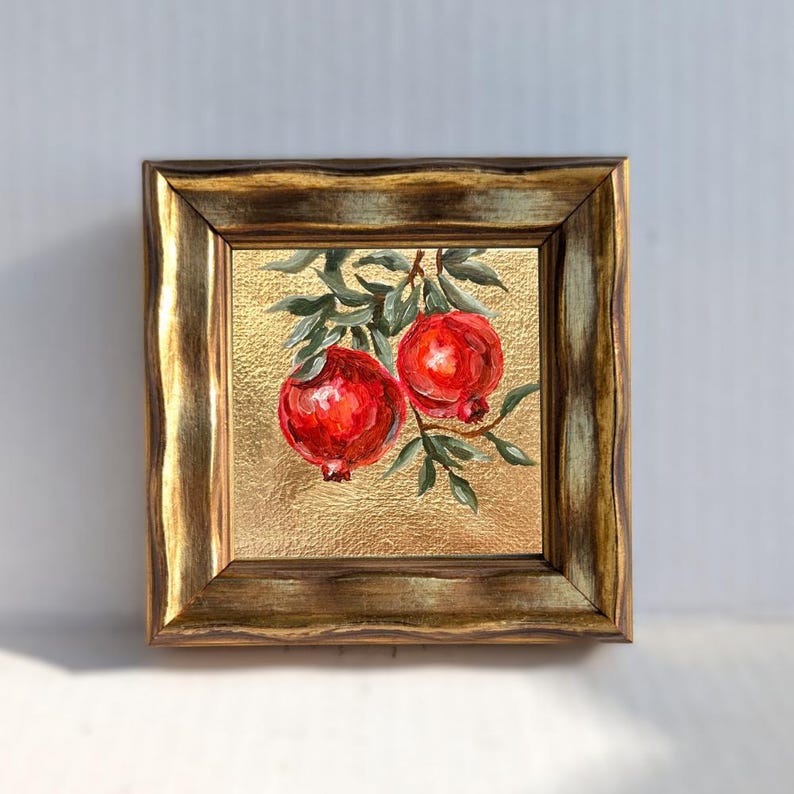 May include: Square framed painting featuring two vibrant red pomegranates on a branch with green leaves. The background is a textured gold color, and the frame is a dark, ornate gold. A decorative piece for home decor.