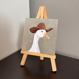 May include: A small painting of a white goose wearing a brown cowboy hat, displayed on a miniature wooden easel. The goose has an orange beak and is set against a muted gray background. The artwork is a whimsical depiction of farm life.