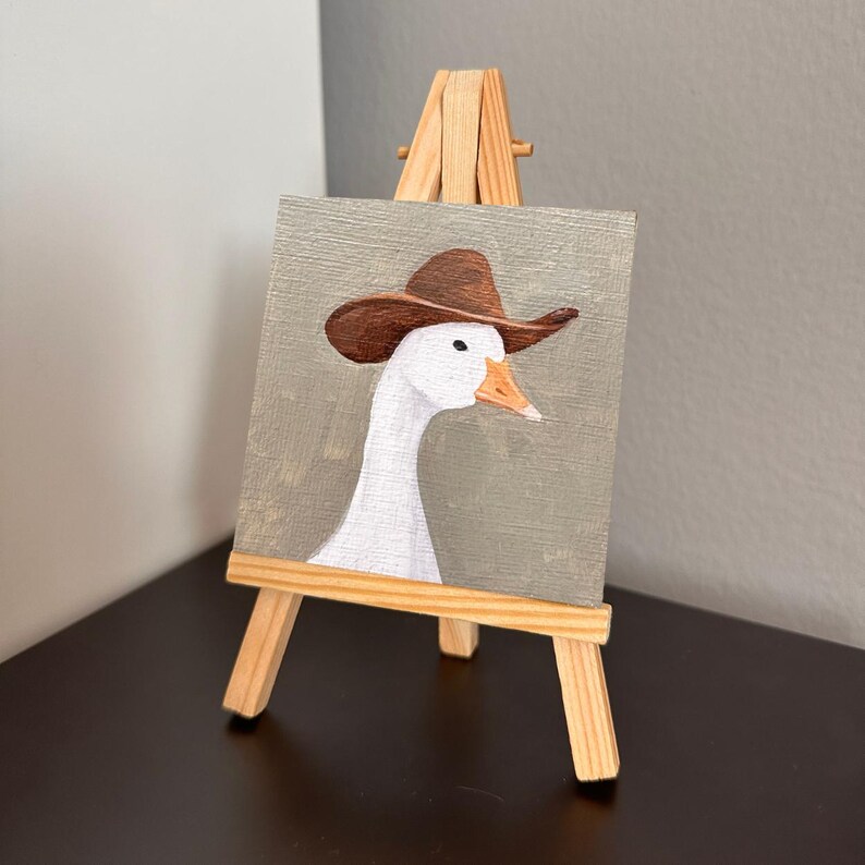 May include: A small painting of a white goose wearing a brown cowboy hat, displayed on a miniature wooden easel. The goose has an orange beak and is set against a muted gray background. The artwork is a whimsical depiction of farm life.