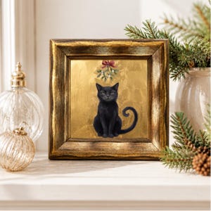 May include: A square framed wall art piece featuring a black cat sitting centered against a metallic gold background. Above the cat, a sprig of mistletoe with a red bow is depicted. The frame is dark, textured, and has a metallic gold finish.