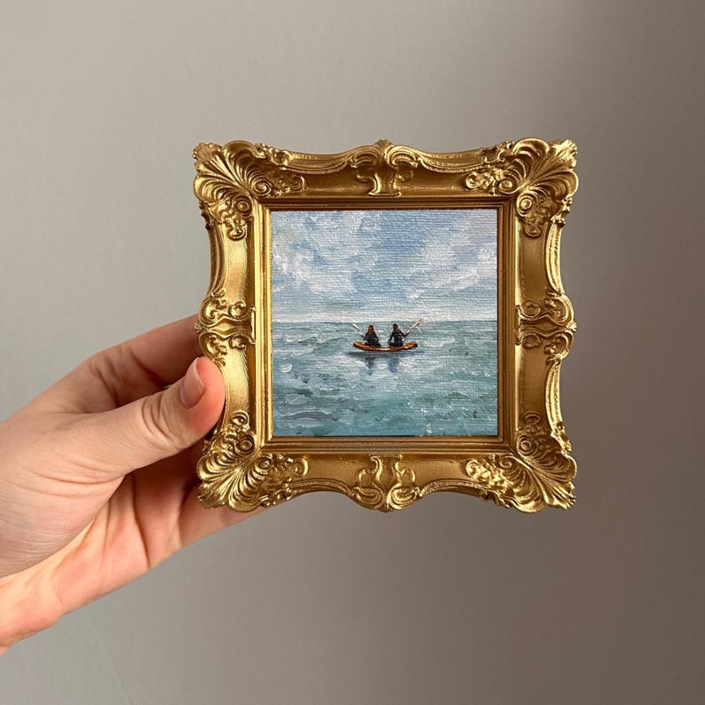 May include: A small, square painting of two figures in a boat on the water, set in a gold ornate frame. The painting depicts a seascape with a blue sky and water. The frame is held by a hand.
