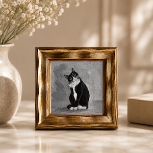 May include: A framed painting of a black and white cat with a gold-colored wooden frame. The cat is sitting and looking to the side. The painting is on a gray background. The frame is square and has a textured appearance.