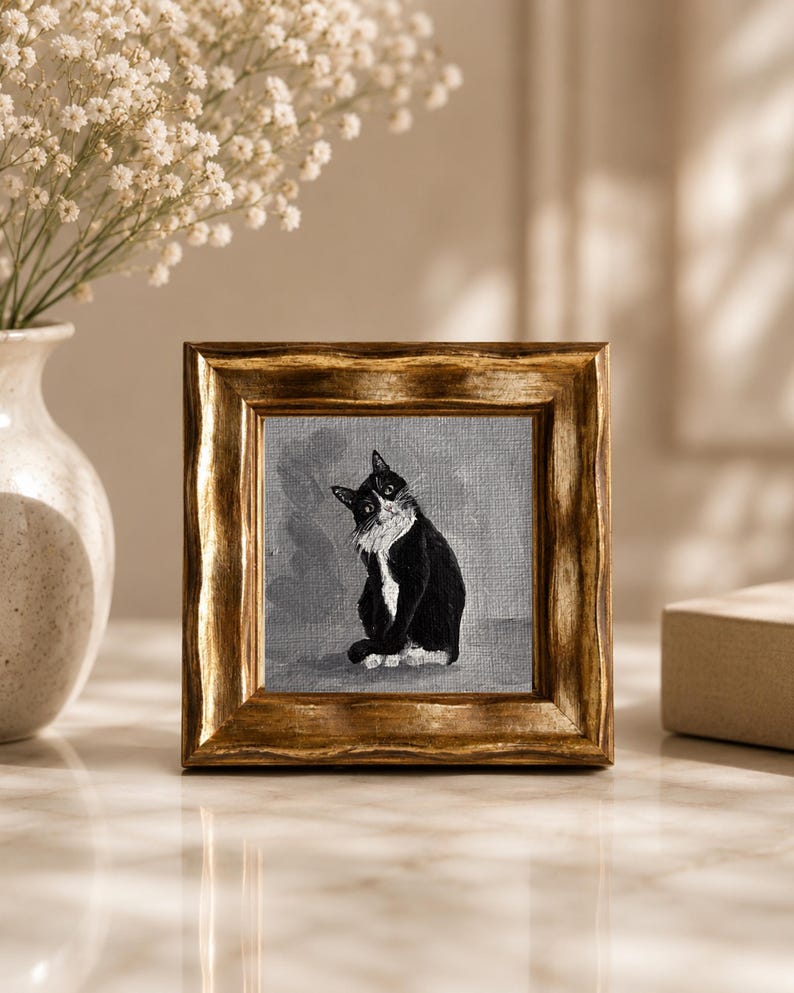 May include: A framed painting of a black and white cat with a gold-colored wooden frame. The cat is sitting and looking to the side. The painting is on a gray background. The frame is square and has a textured appearance.