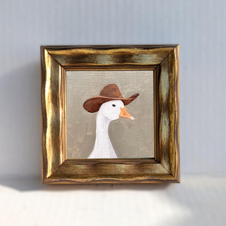 May include: A framed painting of a white goose wearing a brown cowboy hat. The artwork is set against a neutral background and framed in a gold-toned, ornate frame. The goose has an orange beak and is looking to the right.