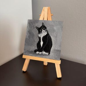 May include: A small, black and white cat portrait on a gray background, displayed on a miniature wooden easel. The cat is painted in a realistic style, with detailed fur and expressive eyes. The artwork is a charming piece of home decor.