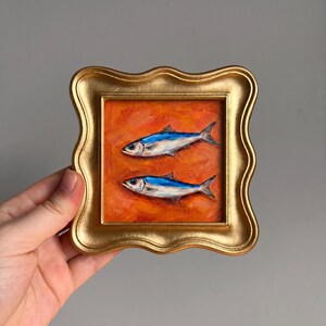 May include: A small, square painting in a gold frame. The artwork features two blue and silver fish on an orange background. The frame has a decorative, wavy edge.