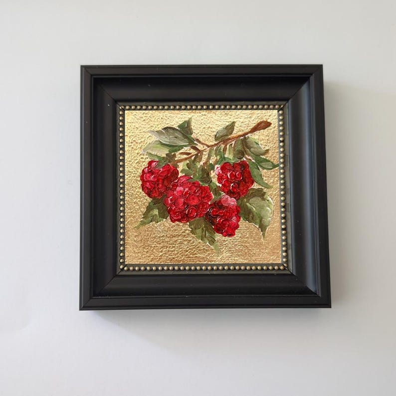 May include: A framed painting of raspberries. The artwork features a branch with vibrant red raspberries and green leaves against a gold textured background. The painting is enclosed in a black frame.