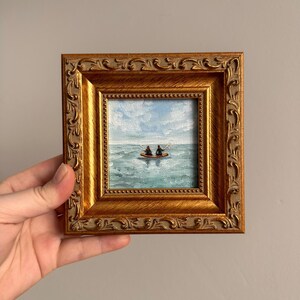 May include: A small, framed painting depicting two figures in a kayak on a turquoise sea under a cloudy blue sky. The painting is set within a gold-colored, ornate frame, adding a touch of elegance to the artwork.