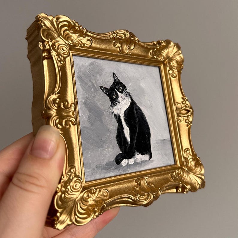 May include: A small, square painting of a black and white cat in a gold ornate frame. The cat is depicted sitting with a curious expression against a gray background. The frame has a detailed, vintage design.