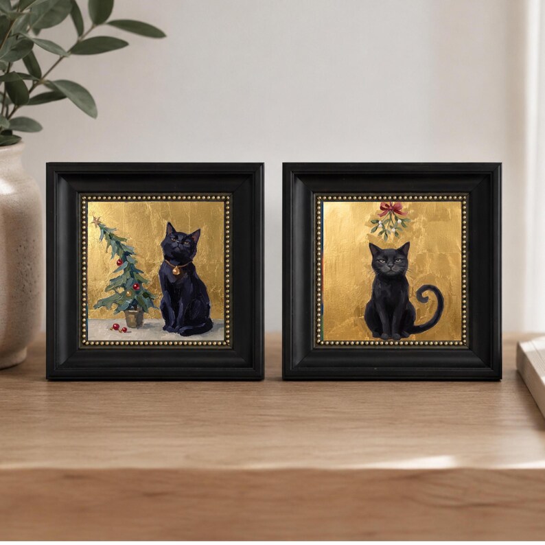 May include: Two square black frames containing paintings of black cats against a gold leaf background. The left painting shows a cat beside a small Christmas tree, while the right painting features a cat sitting beneath a sprig of mistletoe.